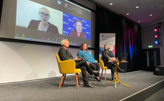 A hybrid event with three people sitting in chairs on a stage and two people on a screen.	